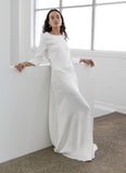 Woman in a long white fashion designer Adrian Hailwood dress standing in a minimalistic room with large windows.